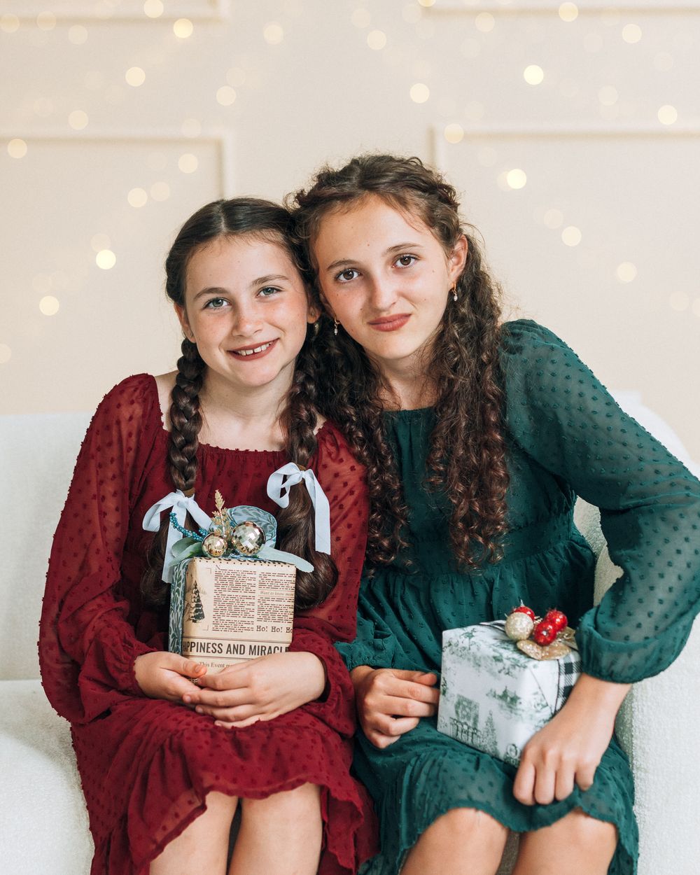 Two girls in festive red and green dresses holding wrapped Christmas gifts, posing together with bokeh lights in background.
