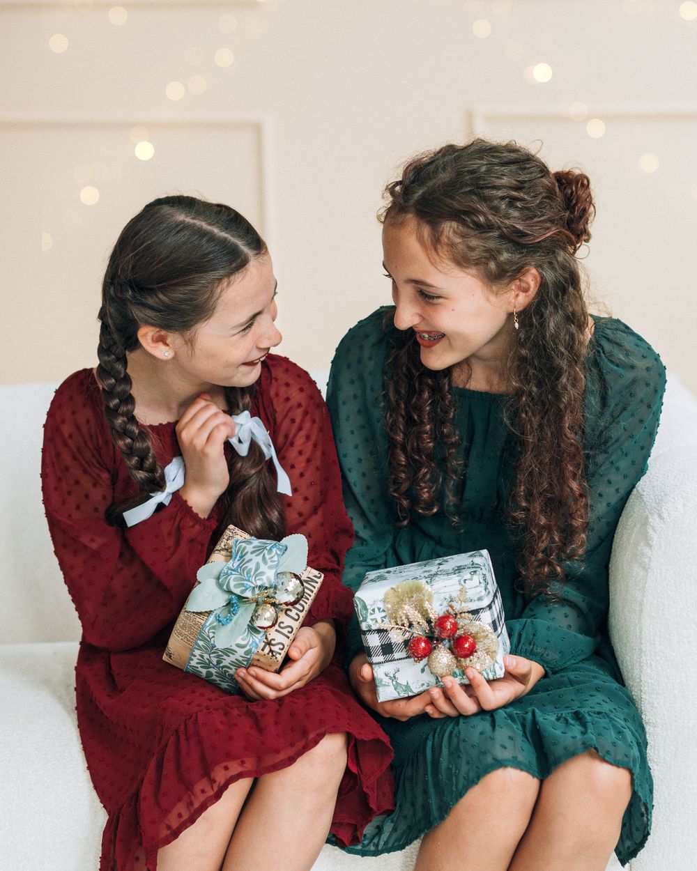 Two girls in red and green dresses smile at each other while holding wrapped Christmas gifts on a white sofa.