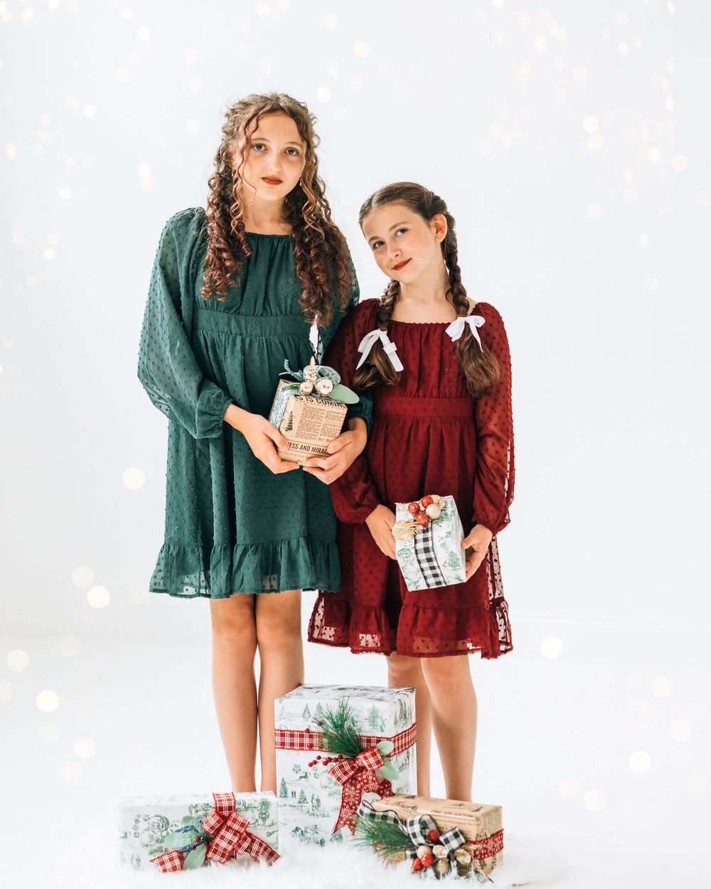 Two girls in festive green and red lace dresses pose with wrapped Christmas gifts on a white background.