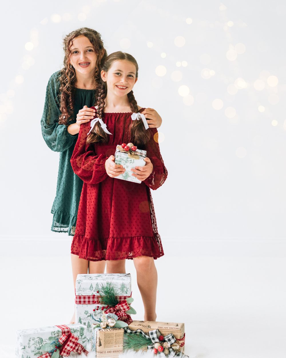 Two girls in festive red and green dresses stand on Christmas gifts with bokeh lights backdrop.