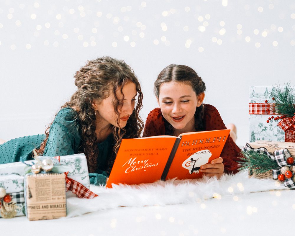 Two girls lying down reading a Merry Christmas book surrounded by festive gifts and bokeh lights.