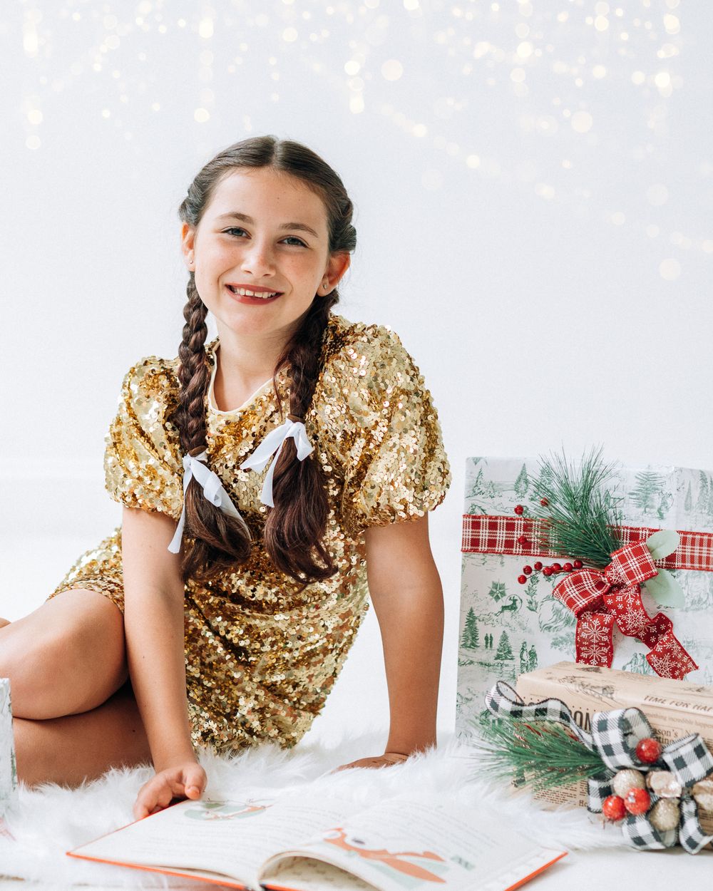 Girl in gold sequin dress with braids sits near Christmas gifts and open book on white fur rug.