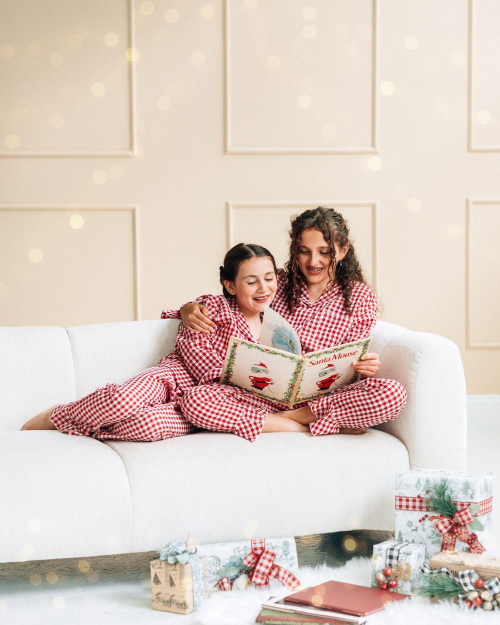 Two girls in matching red gingham Christmas pajamas reading a holiday book together on a white sofa.