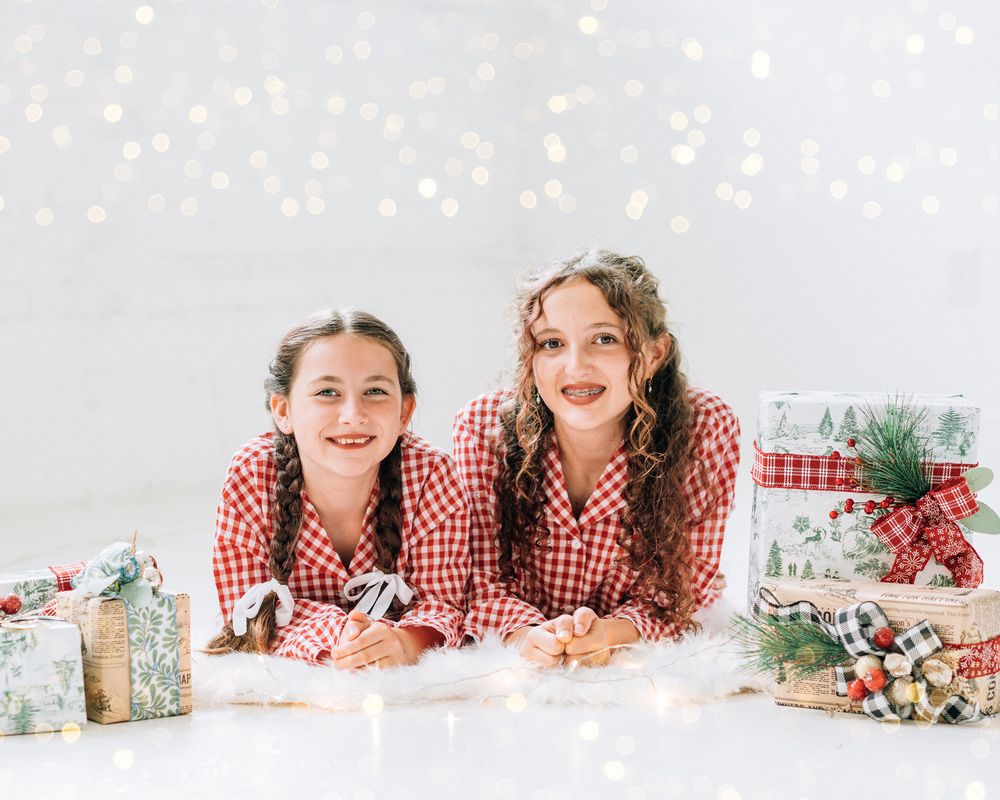 Two smiling girls in red plaid pajamas lying among Christmas gifts with bokeh lights backdrop.