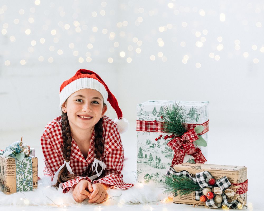 A smiling girl in a Santa hat and plaid pajamas lies among wrapped Christmas gifts with bokeh lights in the background.