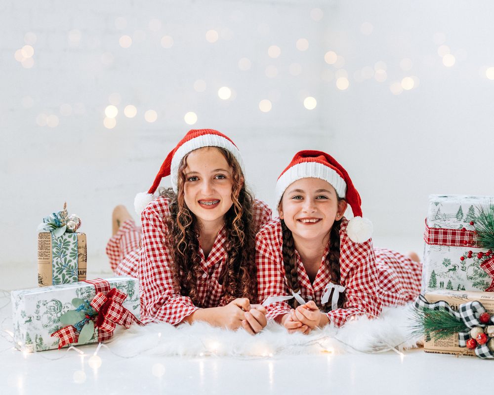 Two girls in red plaid pajamas and Santa hats smile while lying among Christmas gifts with bokeh lights.