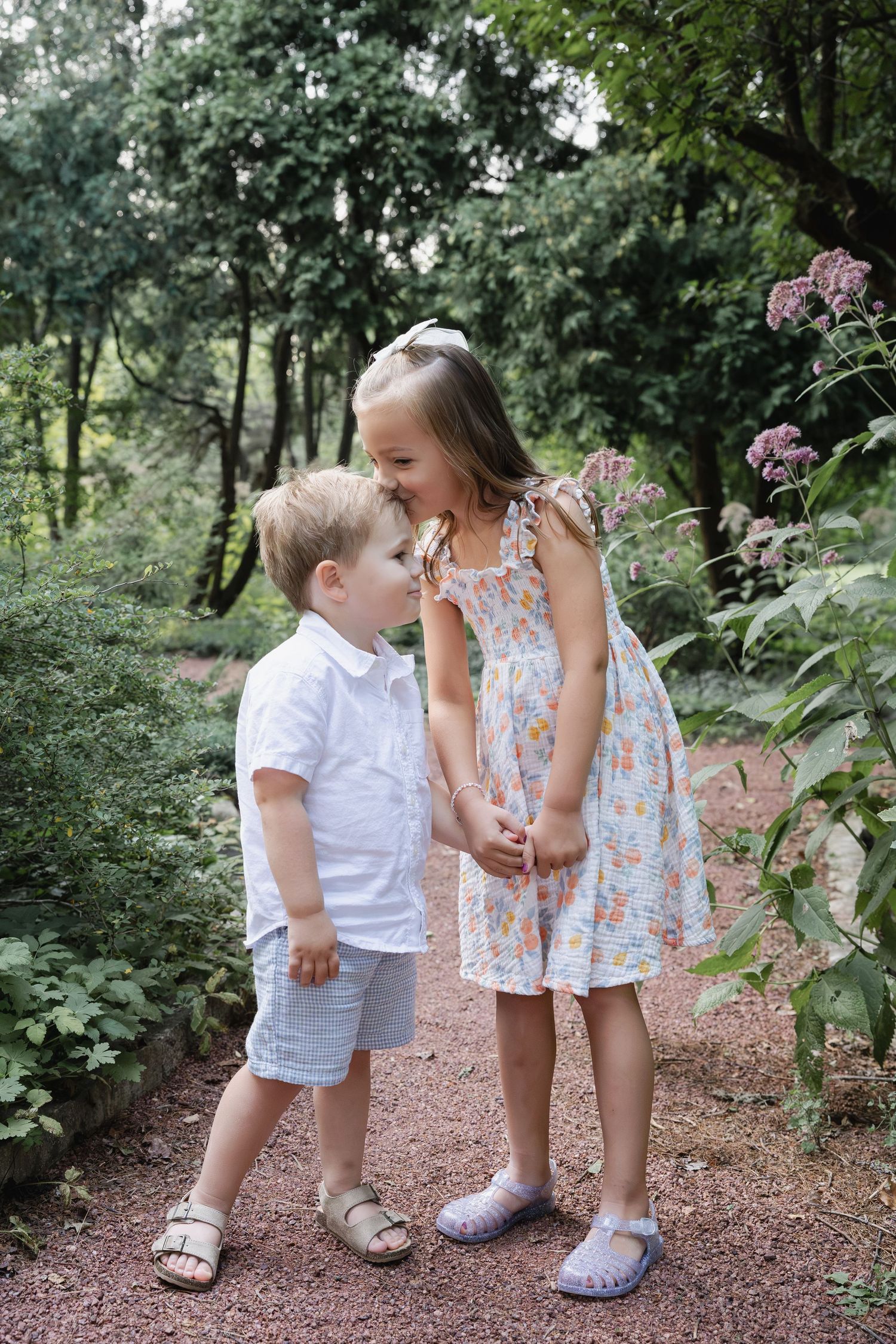 Two young children share a sweet moment on a garden path surrounded by lush green trees and pink flowers.