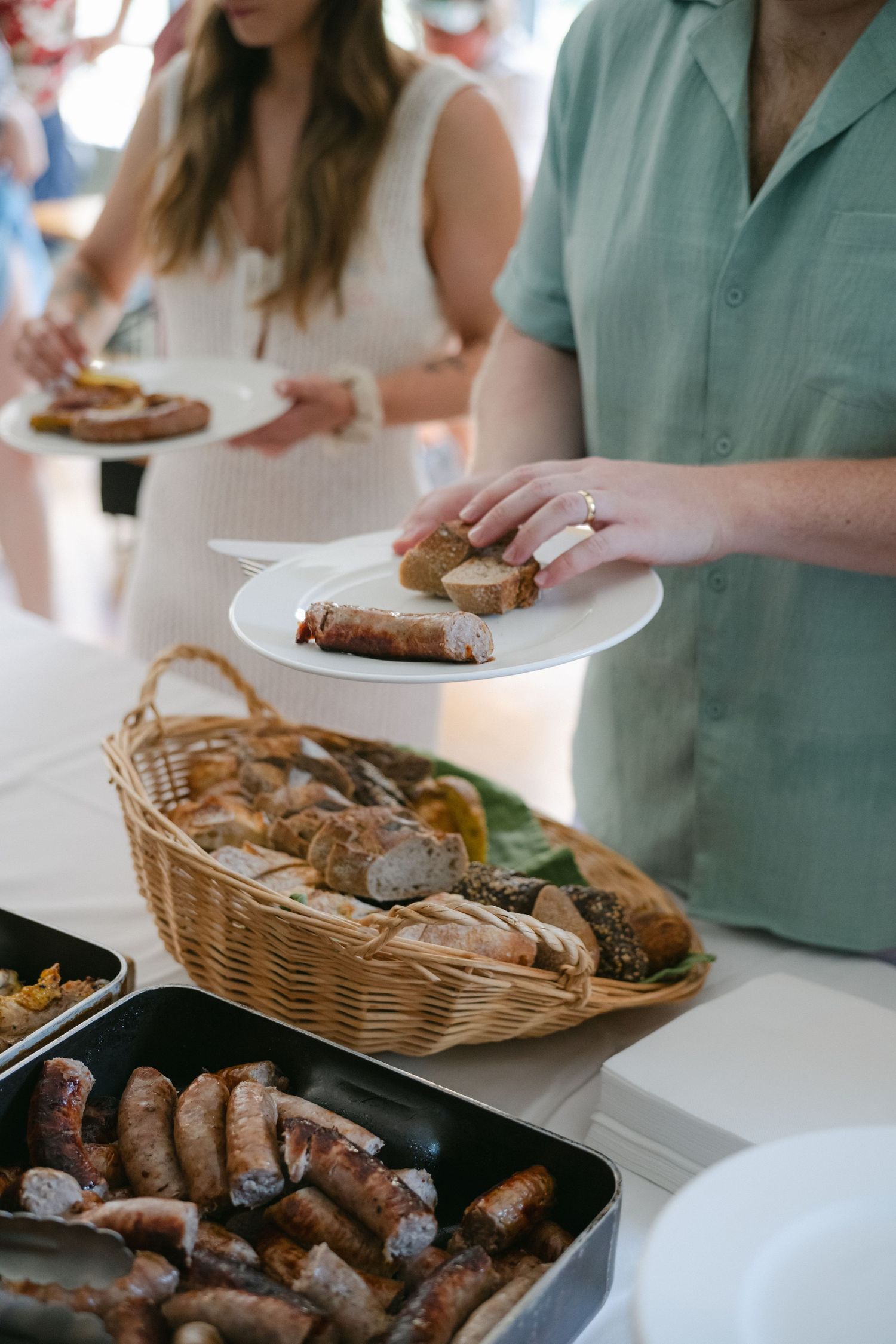 People serving themselves sausages and bread rolls from a wicker basket and trays at a buffet table.
