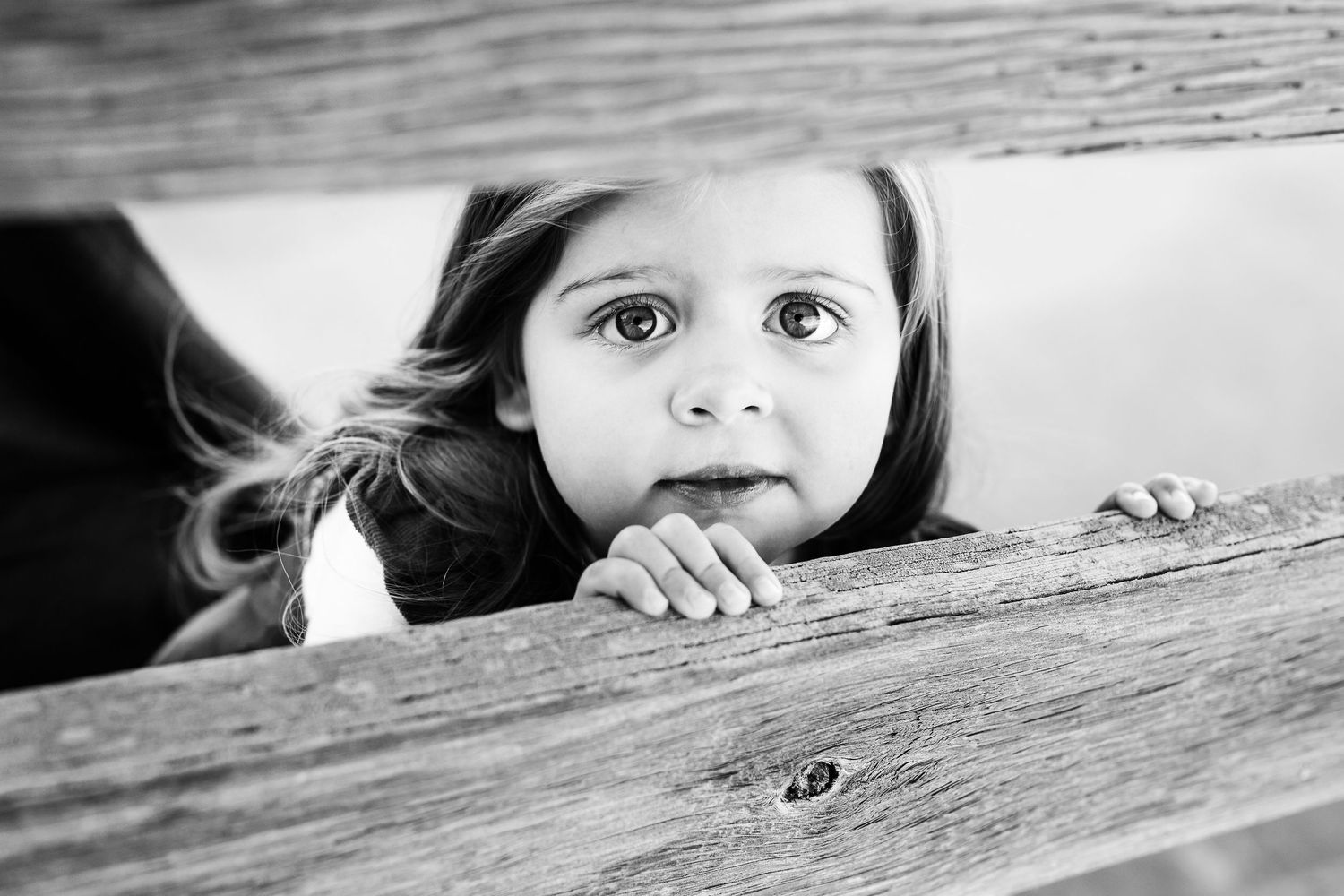 Black and white photo of a curious little girl peeking over a wooden fence with wide, expressive eyes.