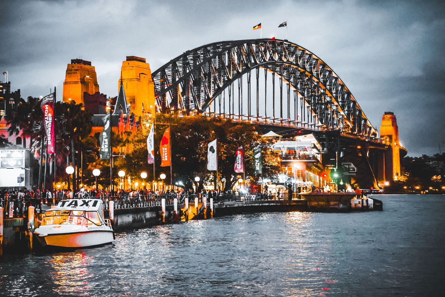Sydney Harbour Bridge illuminated at night with boats and waterfront restaurants reflecting on the water.