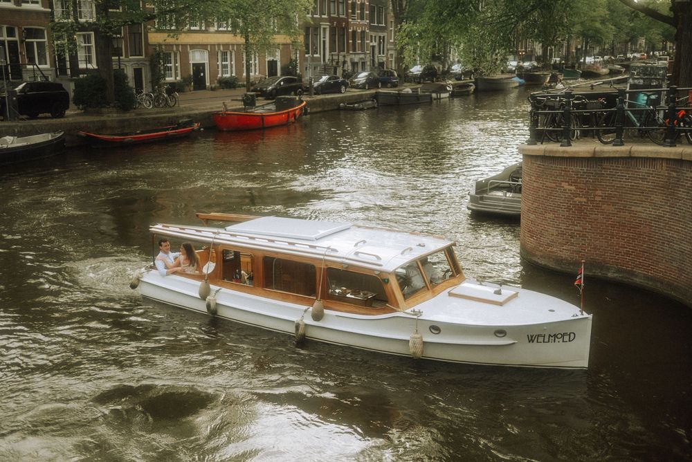 A white canal boat cruising through an Amsterdam waterway, flanked by historic brick buildings and moored boats.