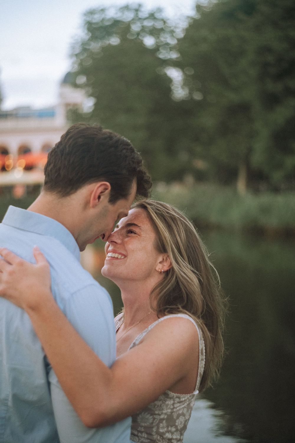 A smiling woman gazes up at a man as they embrace near a serene lake with lush green trees in the background.