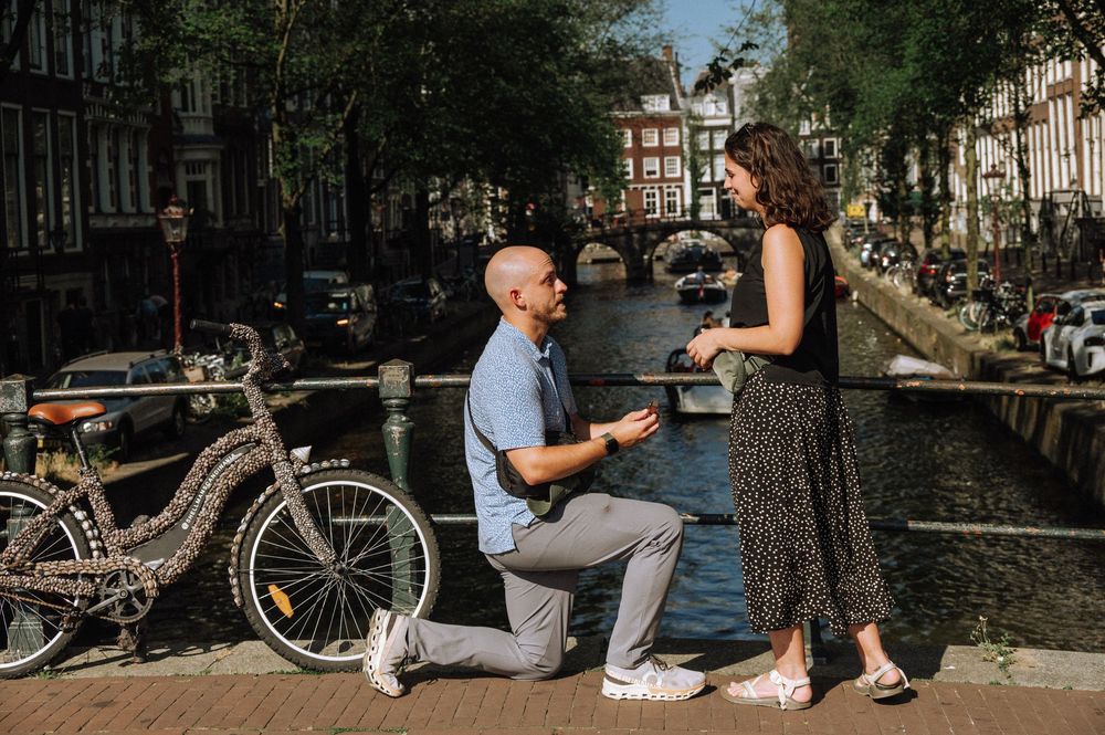 Man proposes to woman on one knee beside Amsterdam canal with bicycle and bridge in background.
