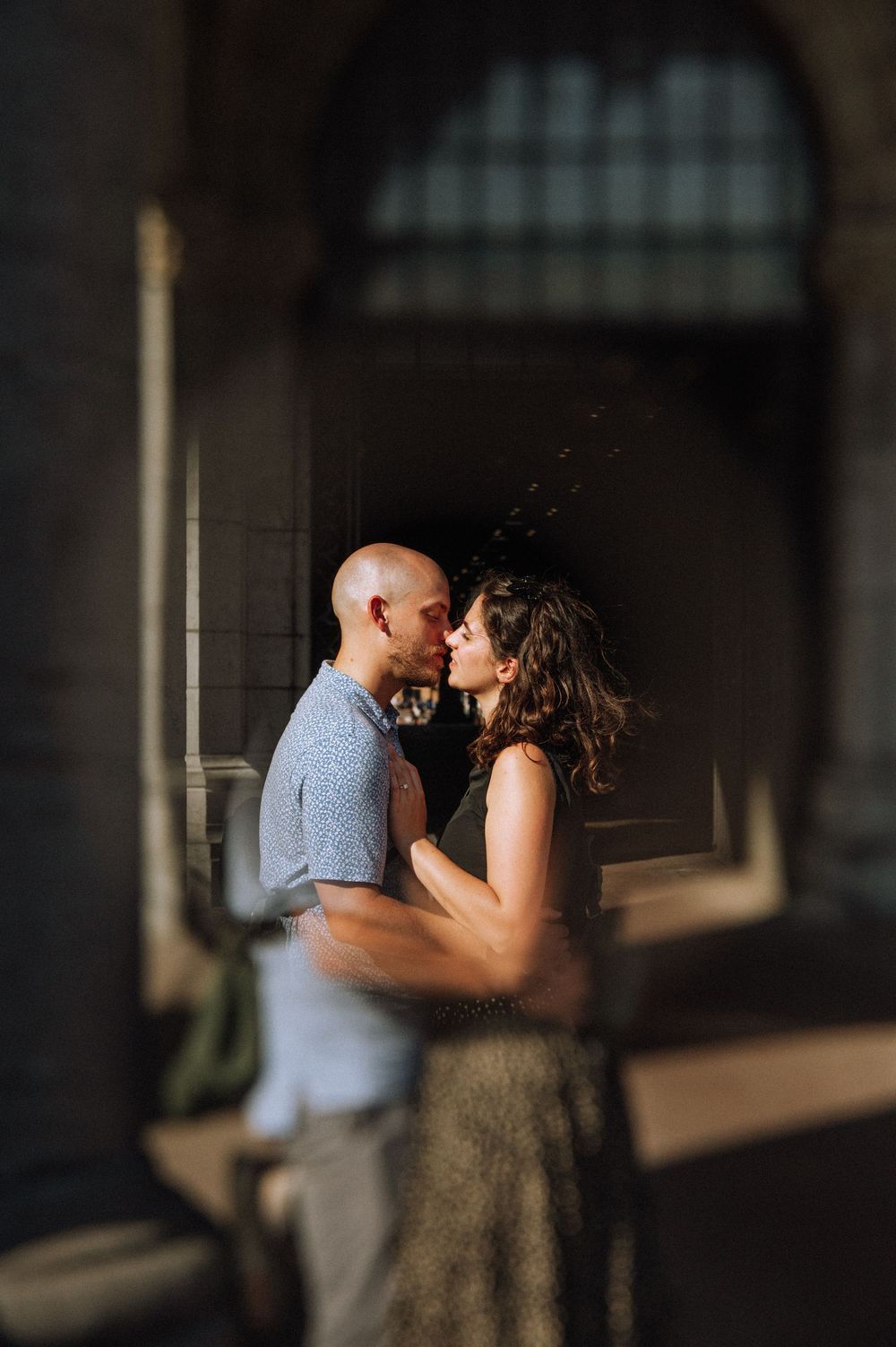 Couple sharing a romantic kiss in a sunlit archway, captured through a dreamy, blurred foreground.