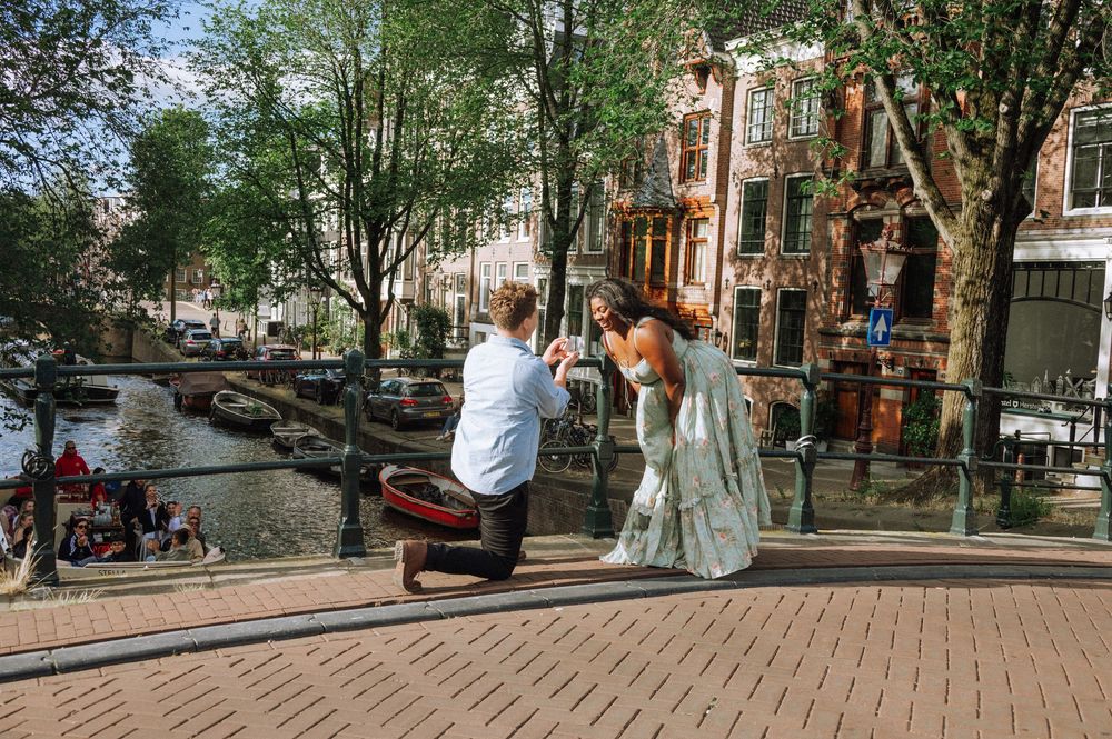 Man proposing to woman on a bridge over Amsterdam canal with historic buildings and trees in background.