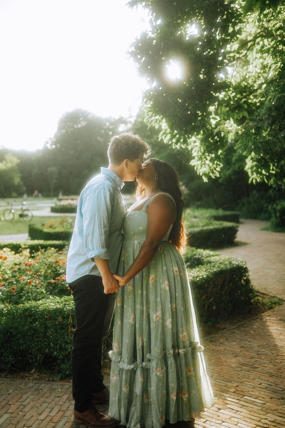 Couple kissing in a sunlit garden park, woman in floral mint dress holding hands with man in blue shirt.