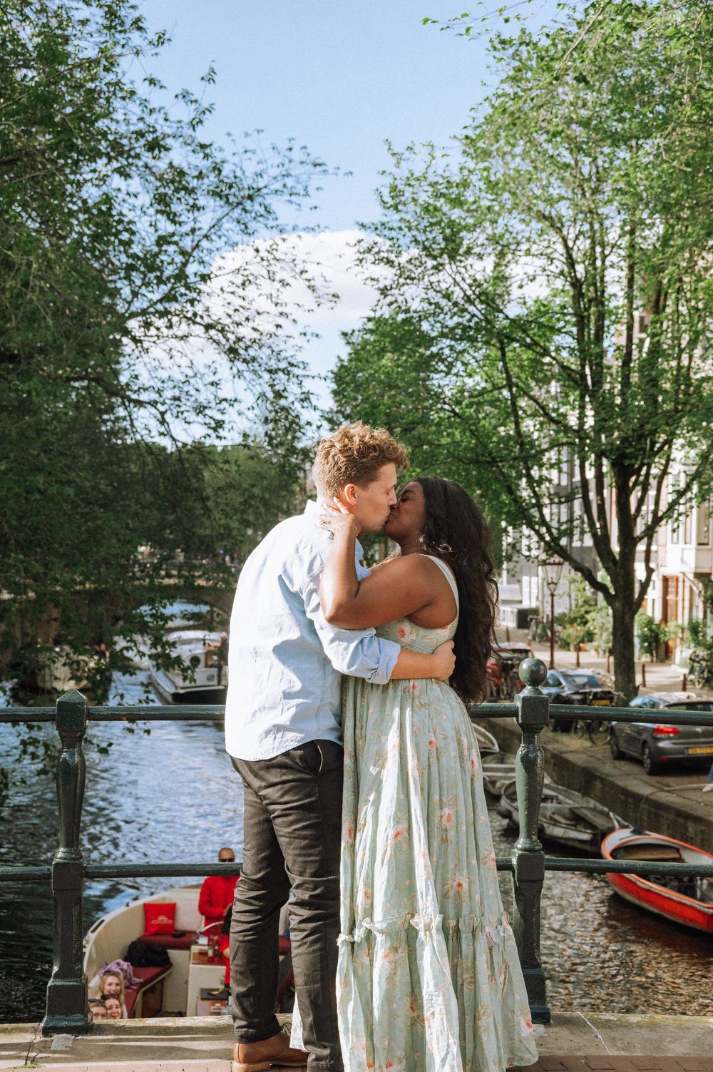A couple sharing a romantic kiss on a canal bridge in Amsterdam, surrounded by lush green trees and boats.