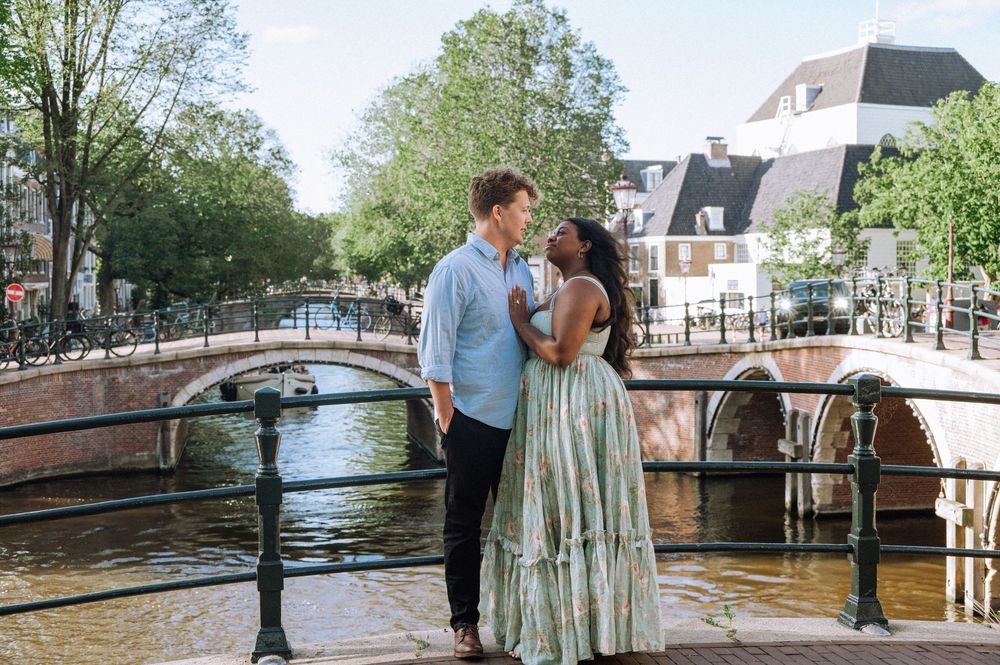 Couple embracing on a canal bridge in Amsterdam, surrounded by lush trees and historic architecture.