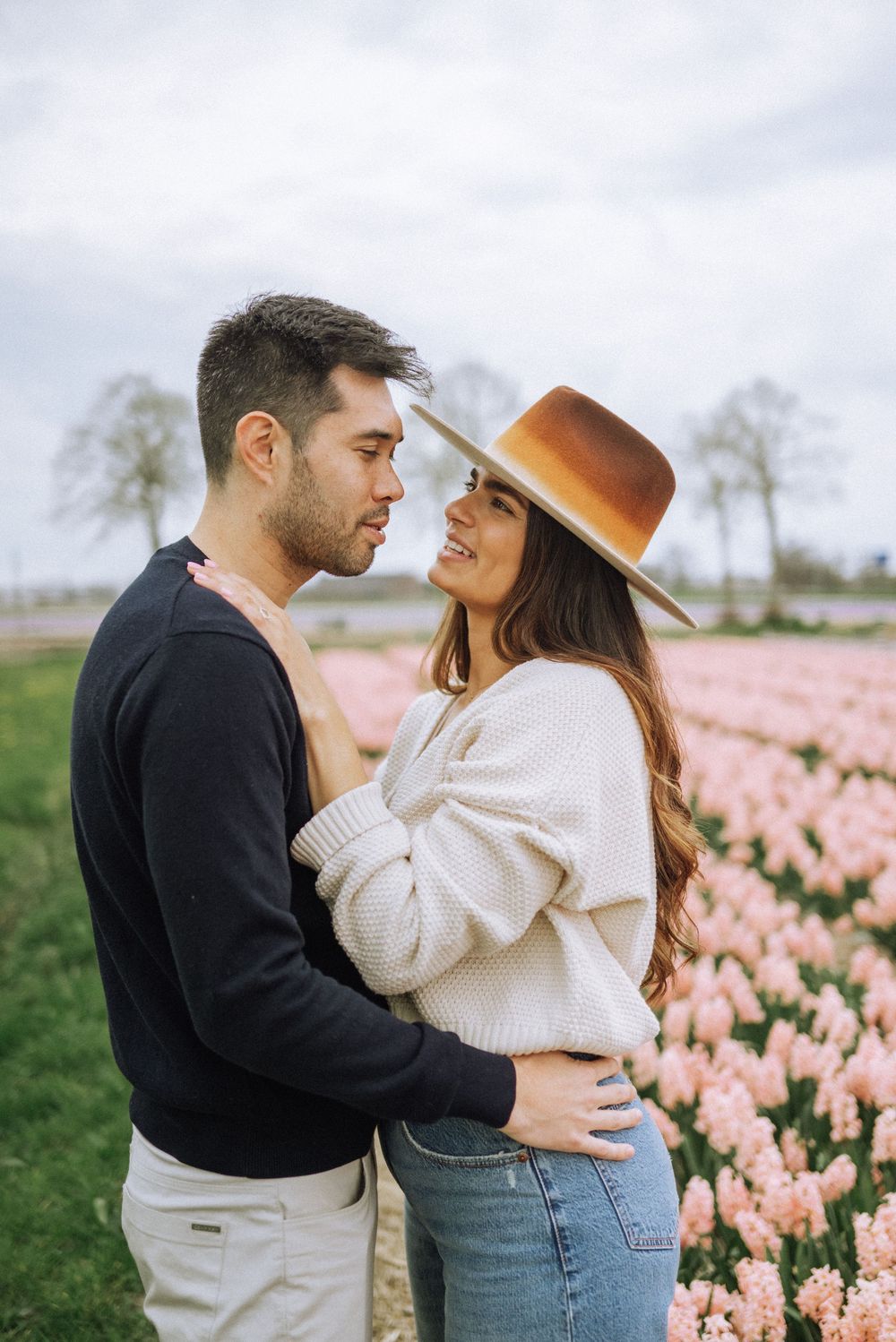 Couple embracing in a pink hyacinth flower field, woman wearing a tan hat and white sweater, man in black sweater.