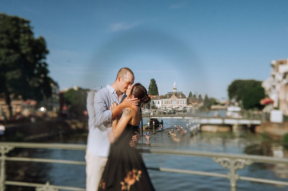 Couple sharing a romantic kiss on a bridge overlooking a scenic waterway on a sunny day.