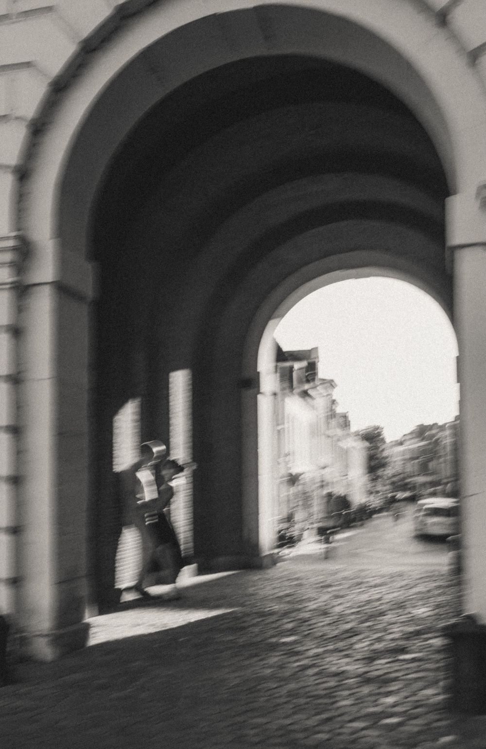 Black and white photo of a person standing under a stone archway, with cobblestone street and buildings visible beyond.