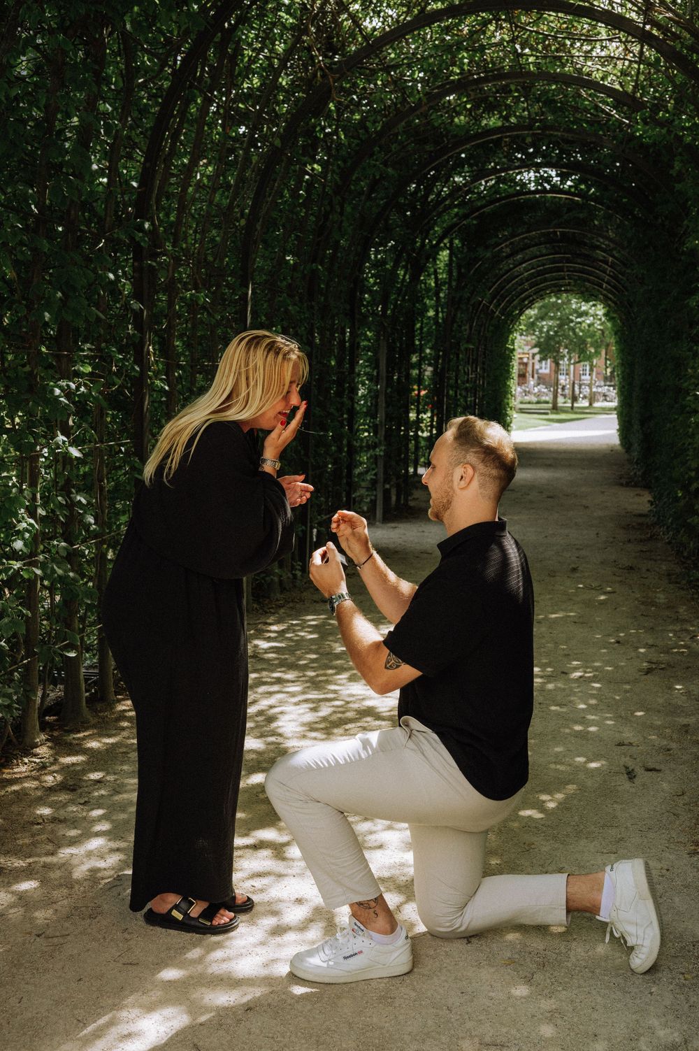 Man proposing on one knee to woman in lush green garden tunnel archway, romantic outdoor marriage proposal.