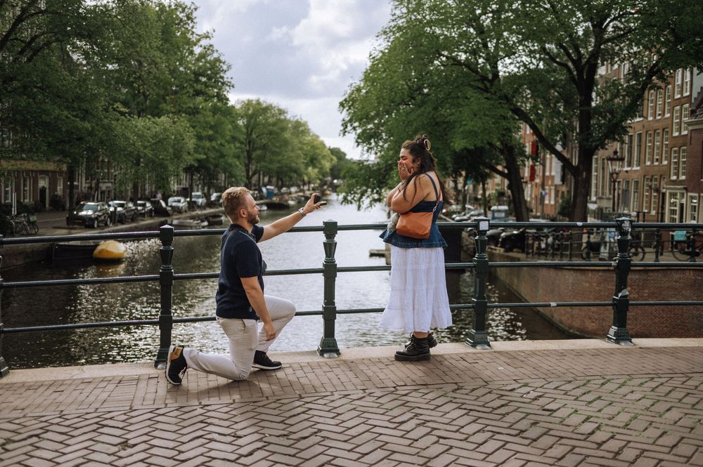 Man proposing to woman on a brick bridge over an Amsterdam canal, surrounded by lush trees and bicycles.