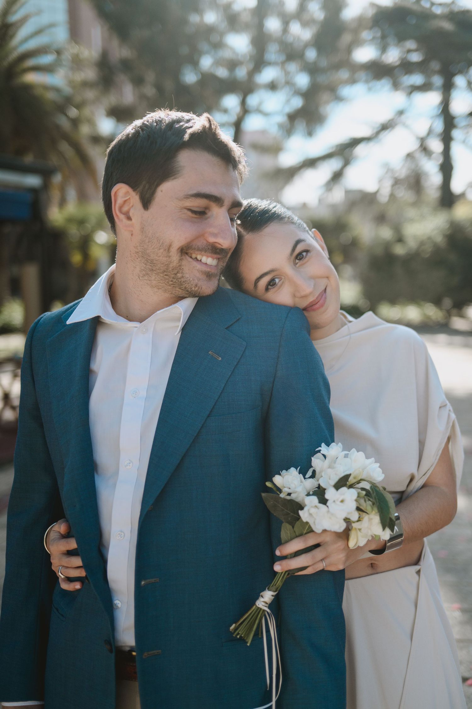 A smiling groom in a blue suit embraces his bride holding white flowers outdoors on their wedding day.