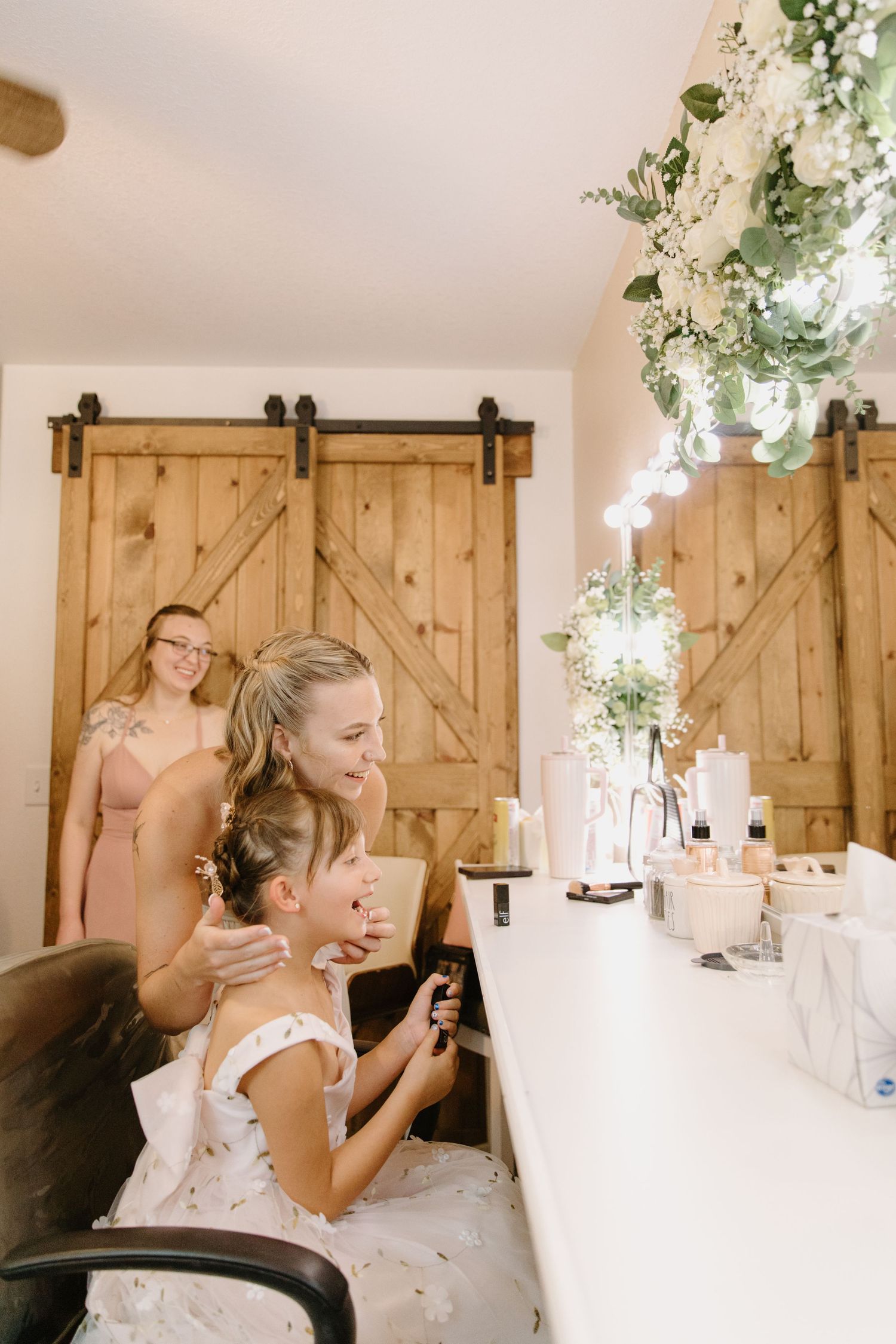 Bride and flower girl share a sweet moment at a vanity mirror in a rustic barn wedding venue with floral decor.