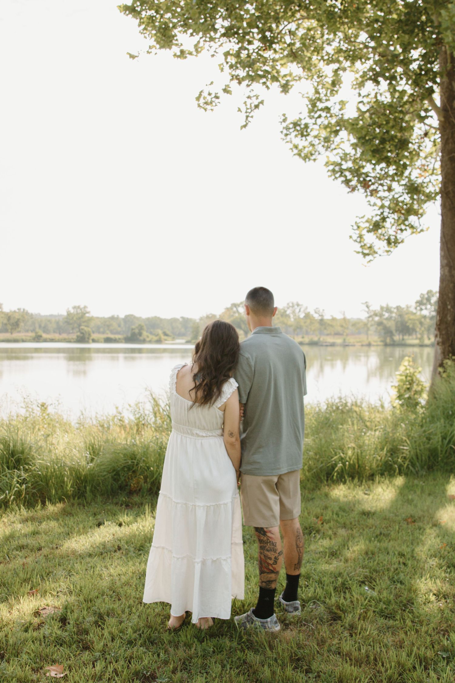 Couple standing together by a serene lake, woman in white dress and man in gray shirt, surrounded by lush greenery.