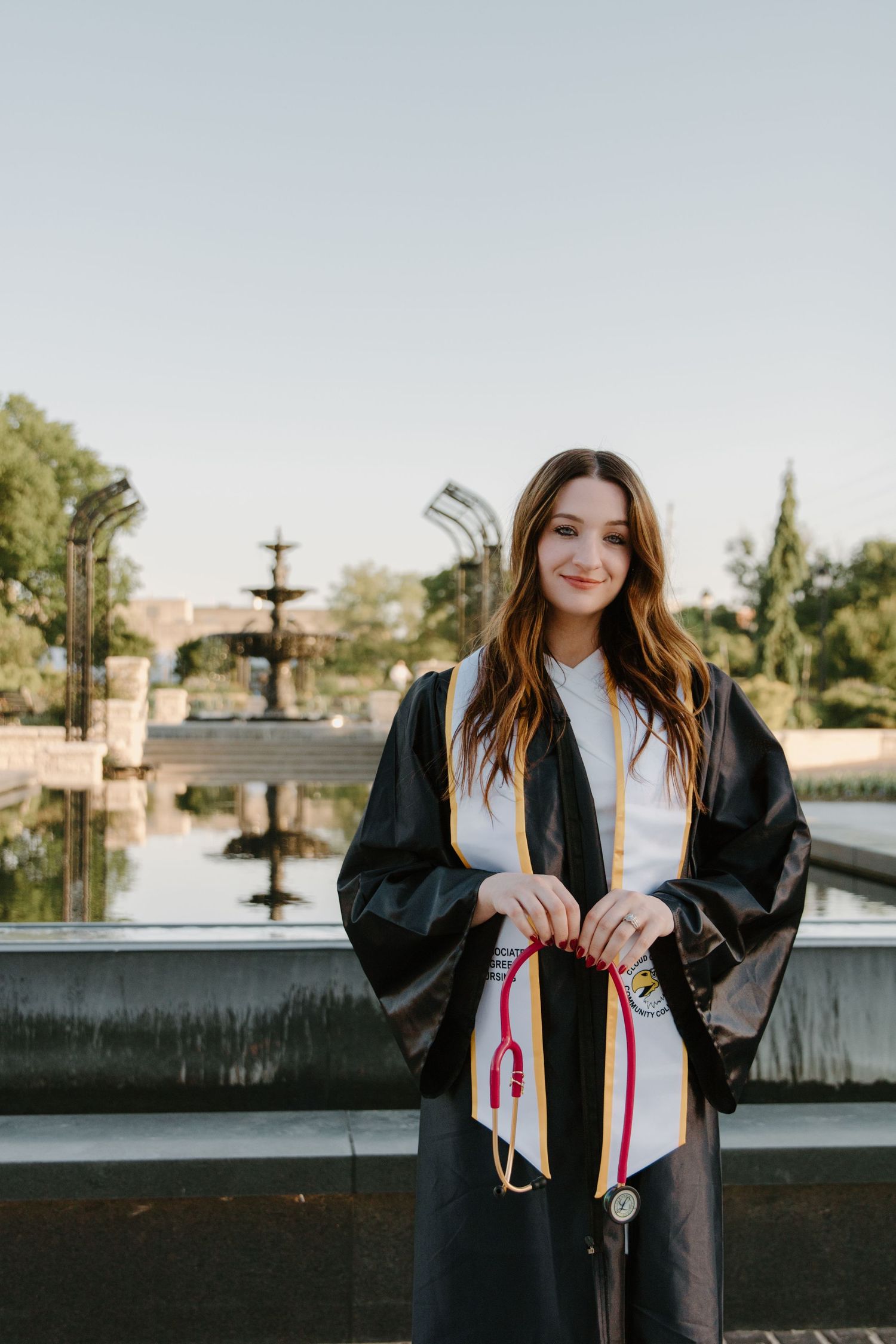 Young woman in black graduation gown holding colorful honor cords, smiling outdoors near a fountain.