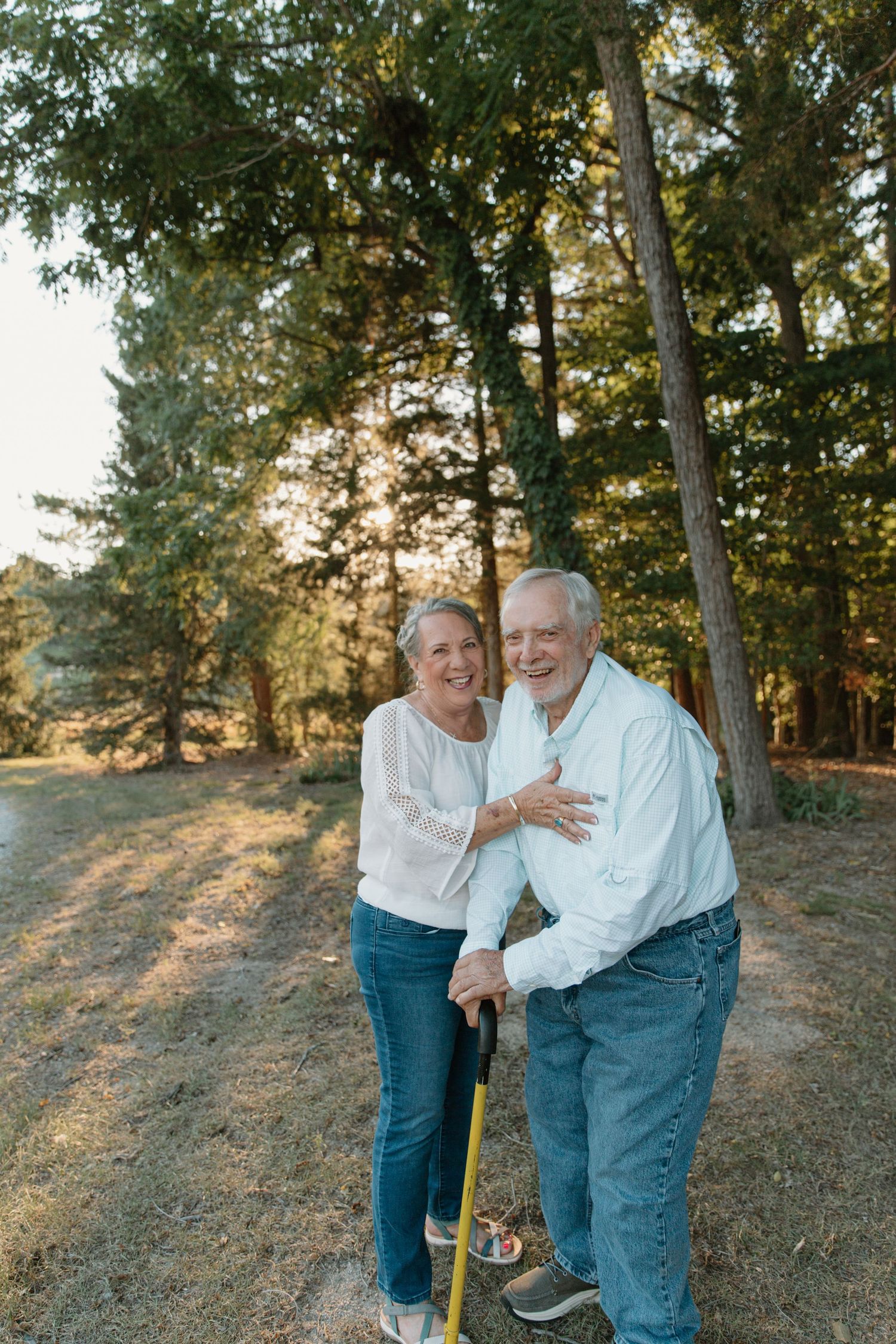 Smiling senior couple in white shirts and jeans pose outdoors among tall green trees in warm golden sunlight.