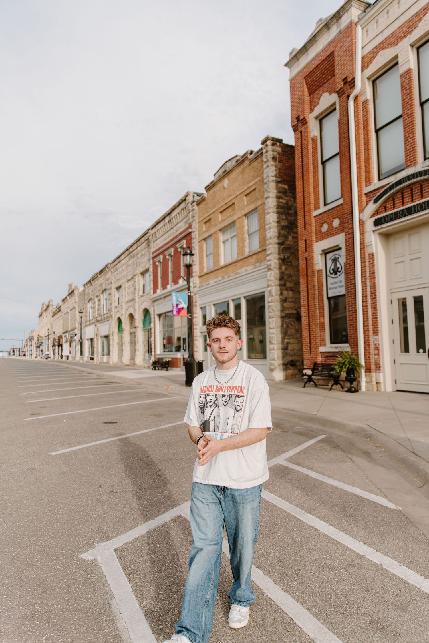 Young man in white graphic tee and jeans standing on empty small town main street with historic brick buildings.