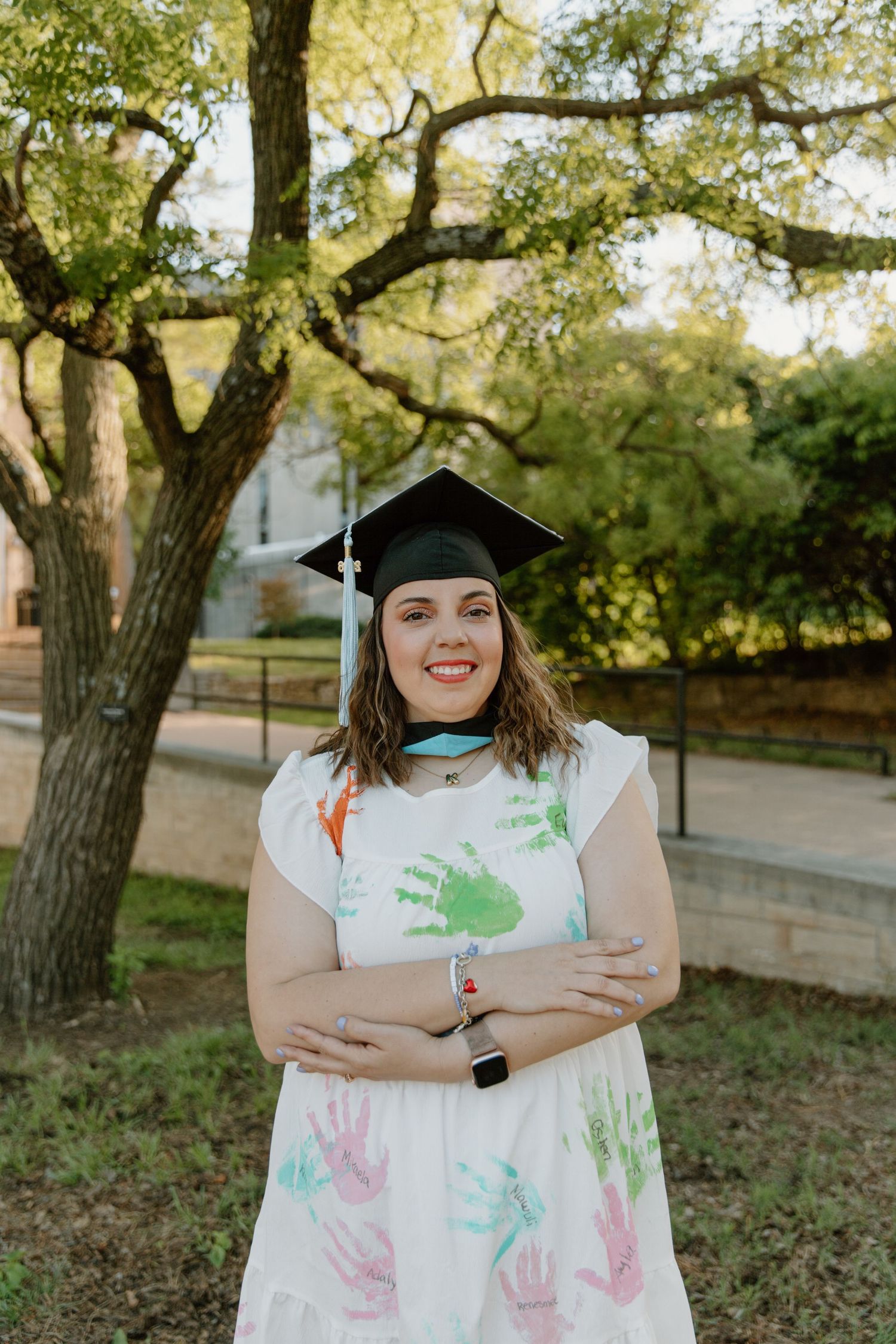 Smiling graduate in cap and handprint dress poses with arms crossed outdoors near trees.