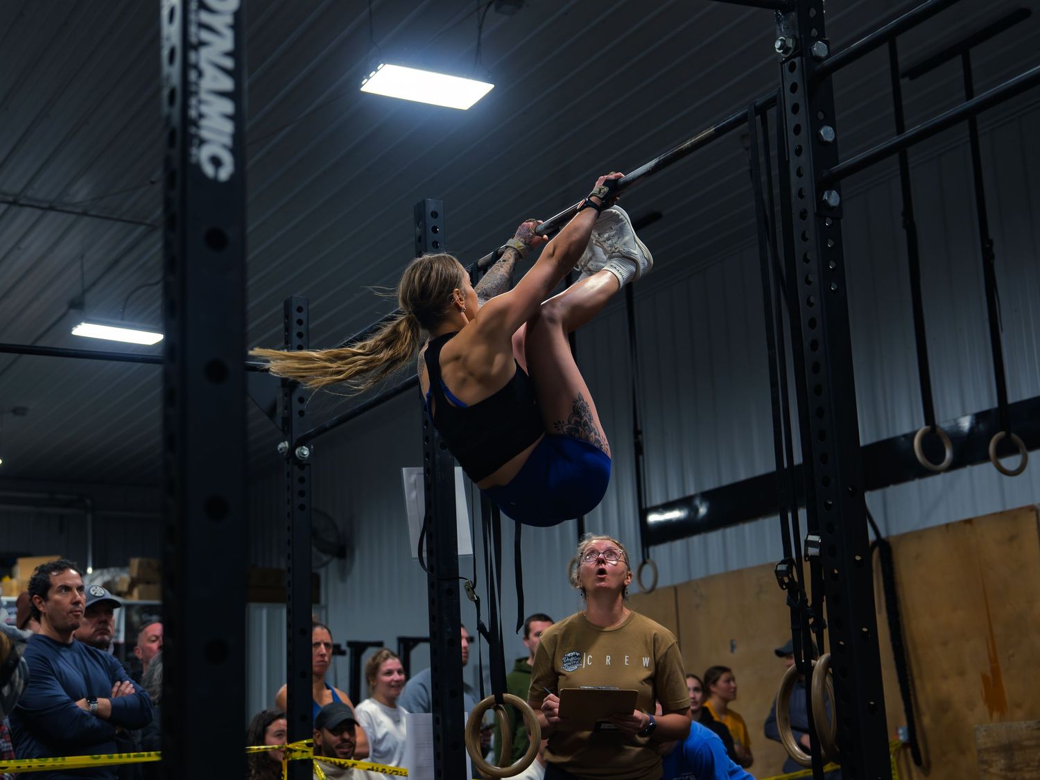 CrossFit athlete drives her toes to the bar on the pull-up rig as a wide-eyed judge tracks the rep below her at a competition.