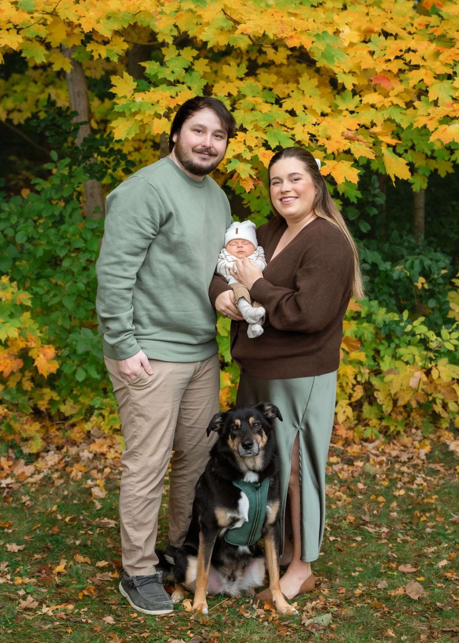Family portrait with newborn baby, dog, and parents in green and brown sweaters, surrounded by vibrant fall foliage.