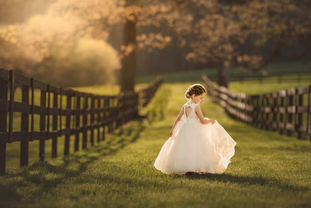 A young girl in a white dress twirls near a wooden fence on a sunlit green lawn at golden hour.