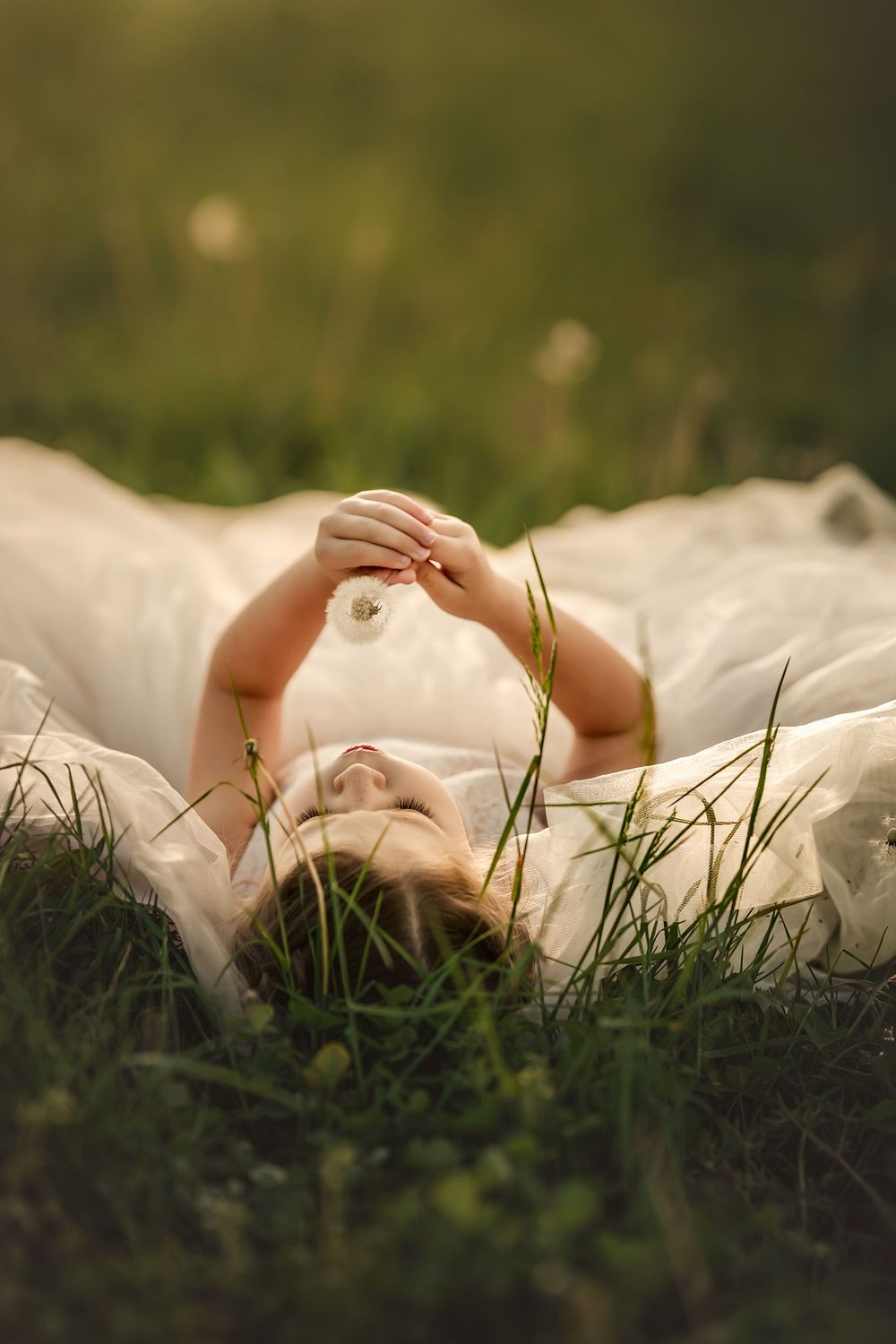 Young girl in white dress lying in green grass, holding a dandelion above her in warm golden sunlight.