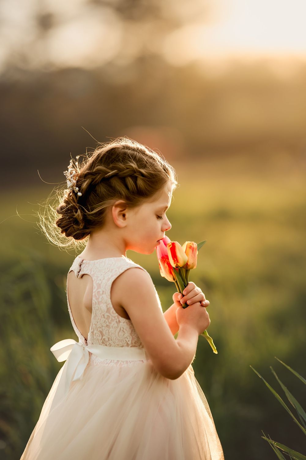 Young girl in white lace dress smelling pink tulips in a sunlit green field.