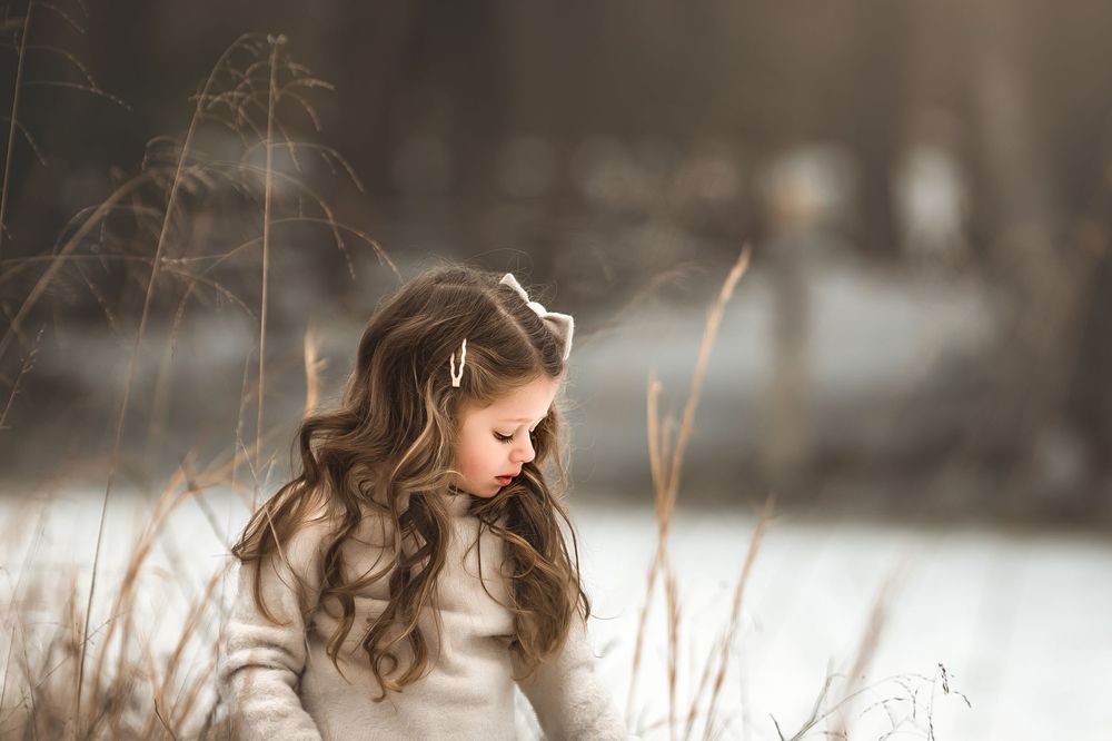 Young girl with long curly hair and hair clip poses outdoors in winter, wearing a white sweater near dry grass.
