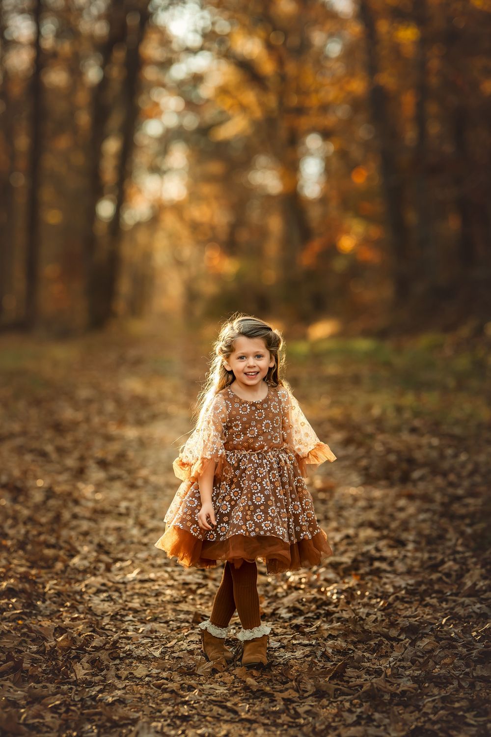 A joyful little girl in a floral brown dress walks along a leaf-covered autumn forest path.