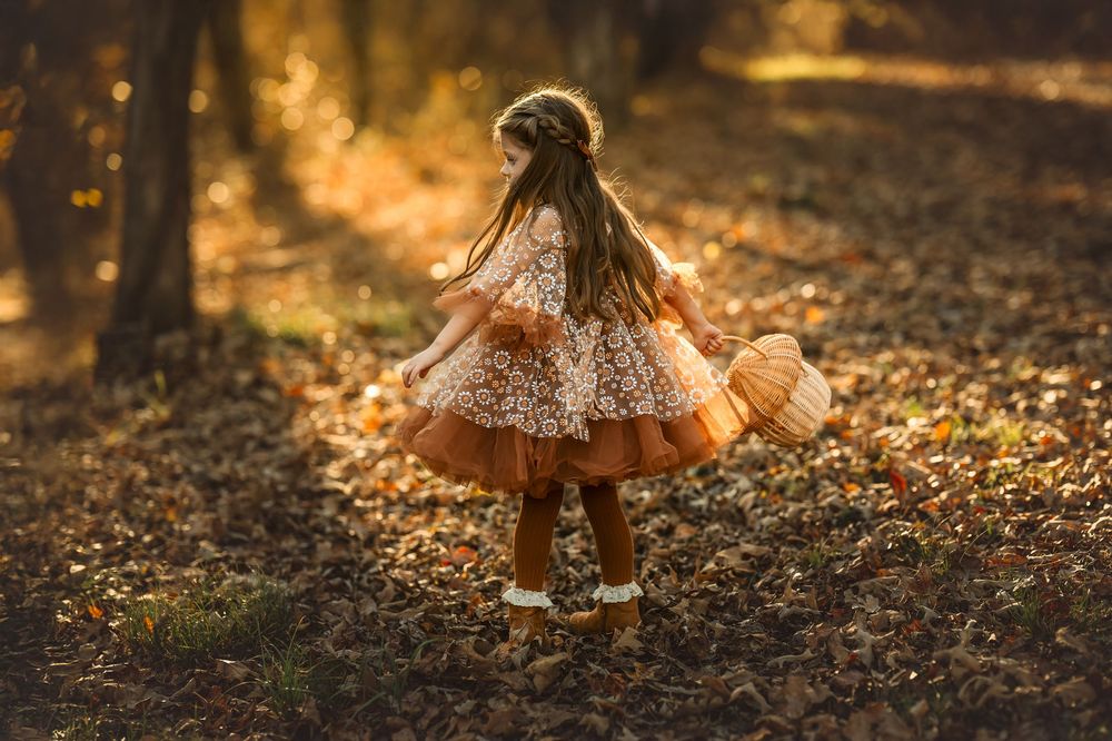 Young girl in floral autumn dress twirling among fallen leaves in golden sunlit forest.