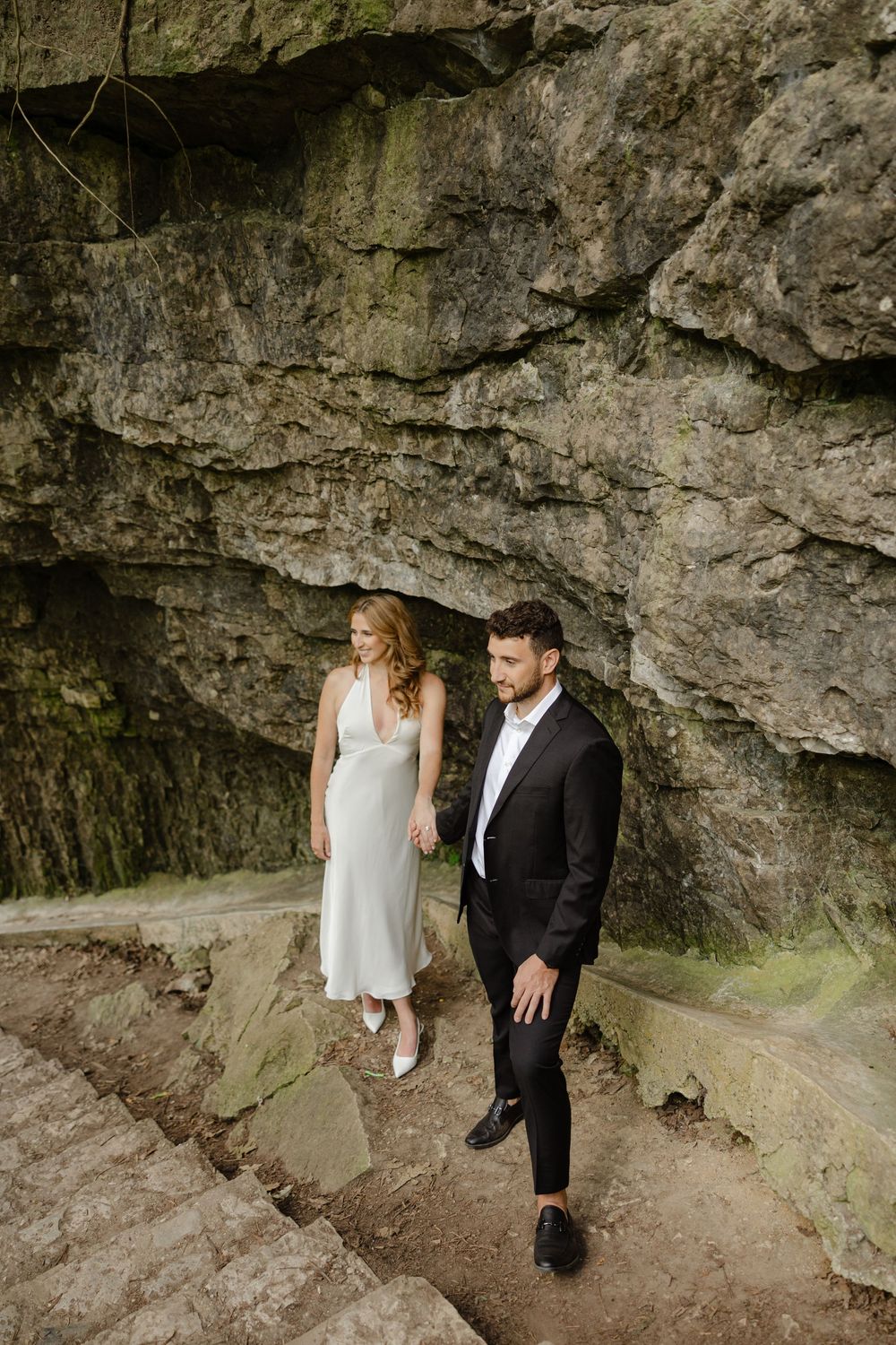 Bride in white halter dress and groom in black suit walking hand in hand along a dramatic rocky cliff face.