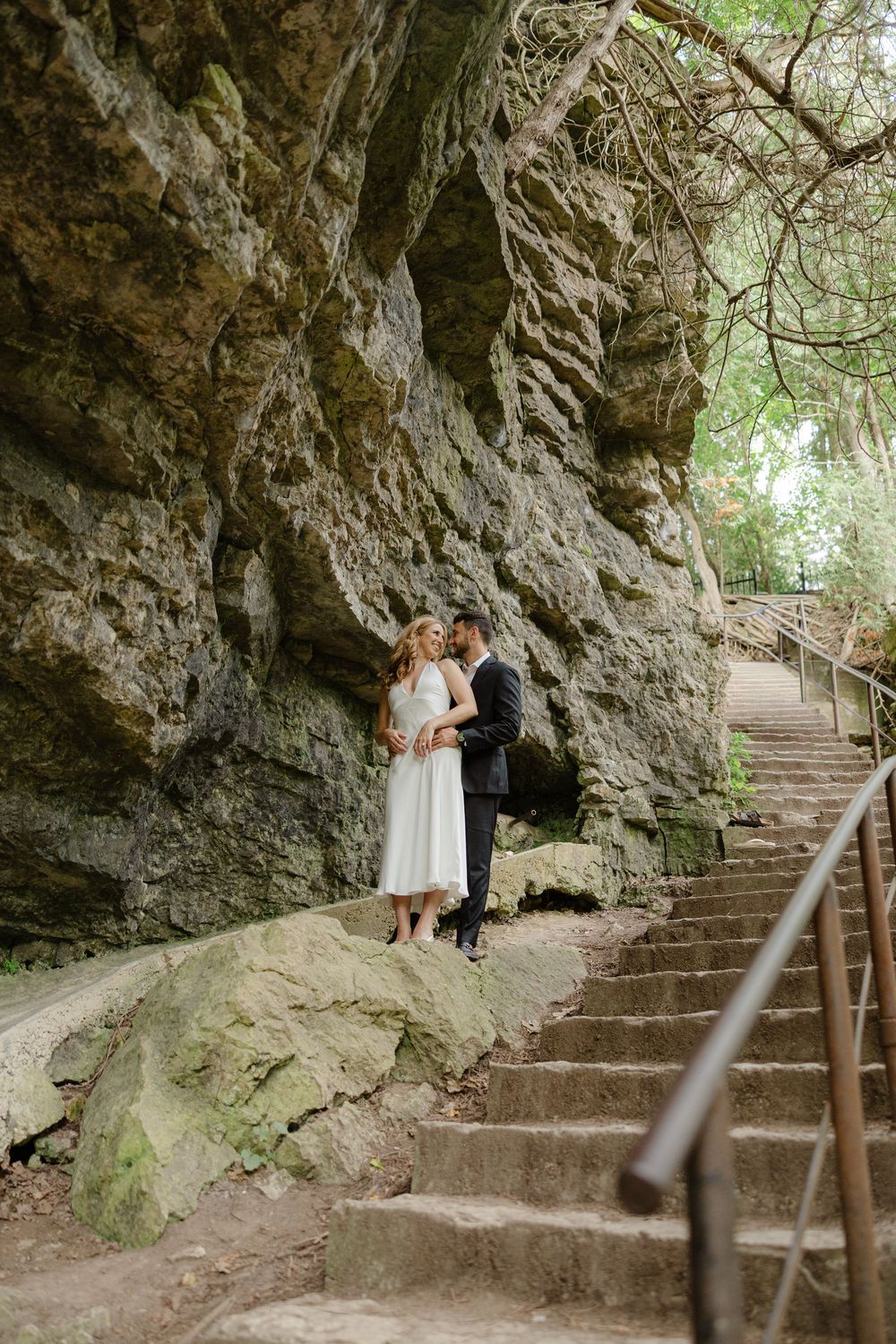 Bride and groom sharing an intimate embrace beside a stunning moss-covered rock face with stone steps nearby.