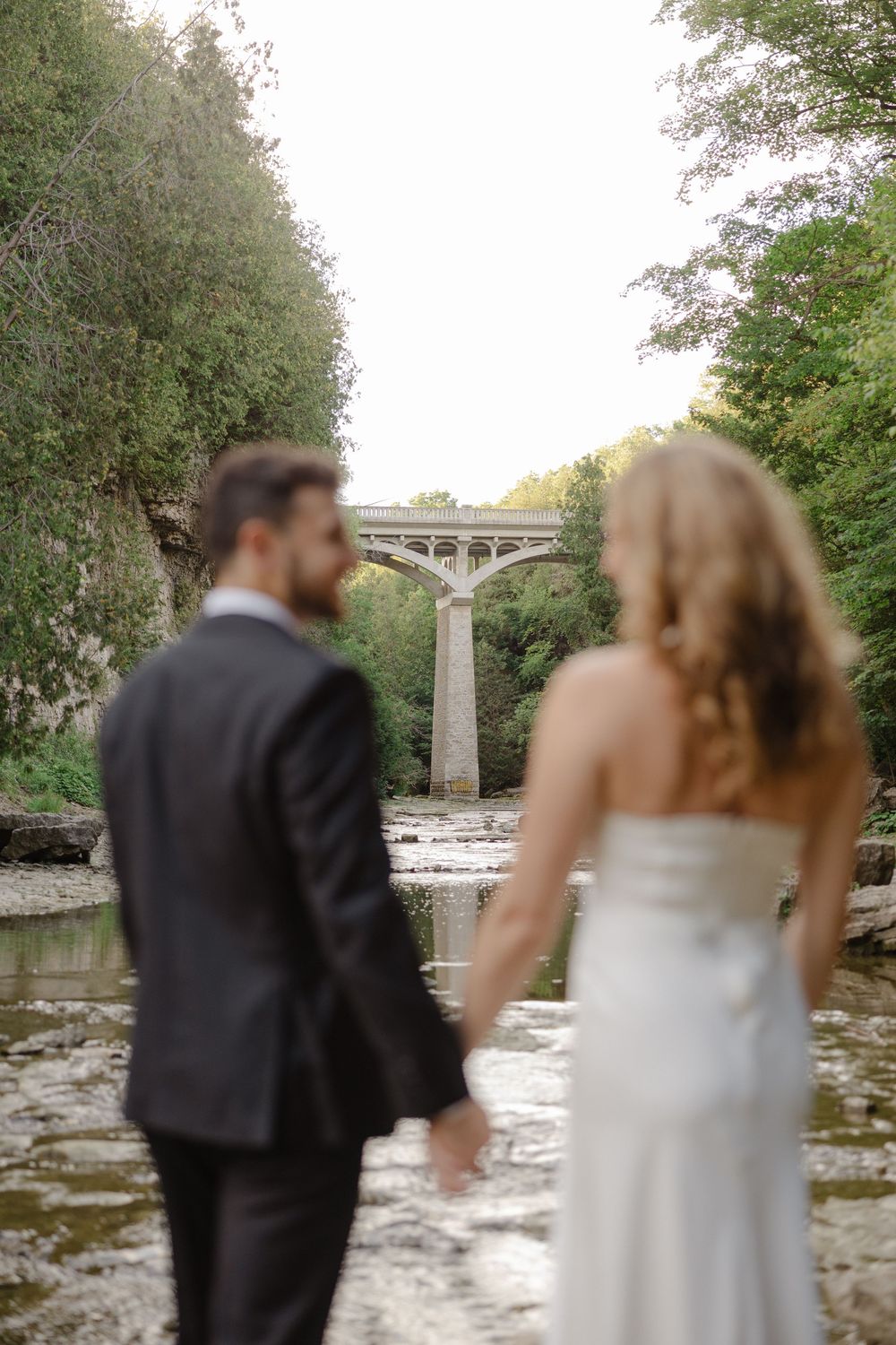 Bride and groom holding hands while gazing at a scenic stone arch bridge over a peaceful river.