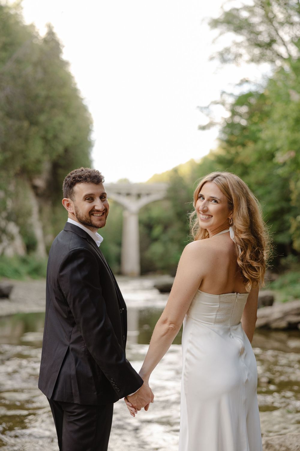 Smiling bride and groom holding hands and looking back at camera with a stone bridge and river backdrop.