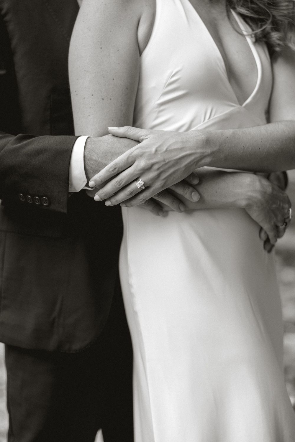 Black and white close-up of groom embracing bride in white satin dress, showing ring.