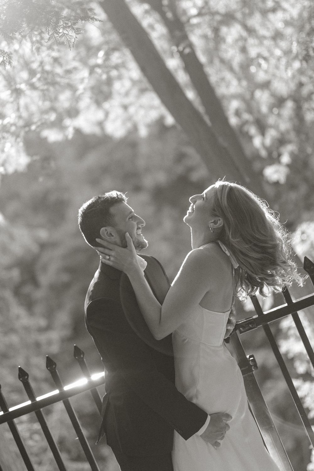 Black and white photo of bride and groom embracing and laughing by an iron fence surrounded by trees.