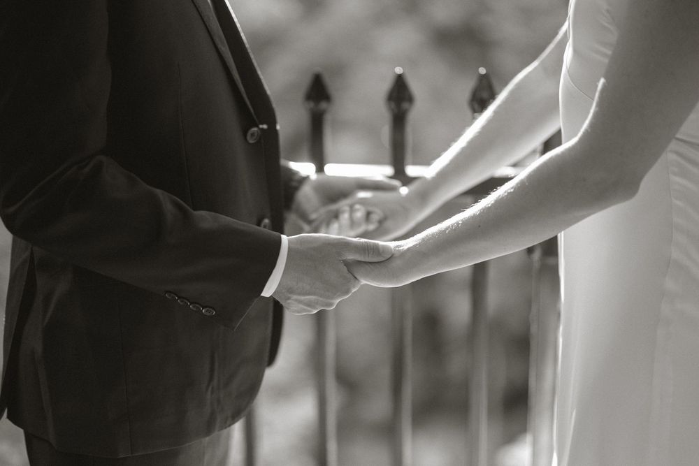Close-up black and white photo of bride and groom holding hands near an ornate iron fence.