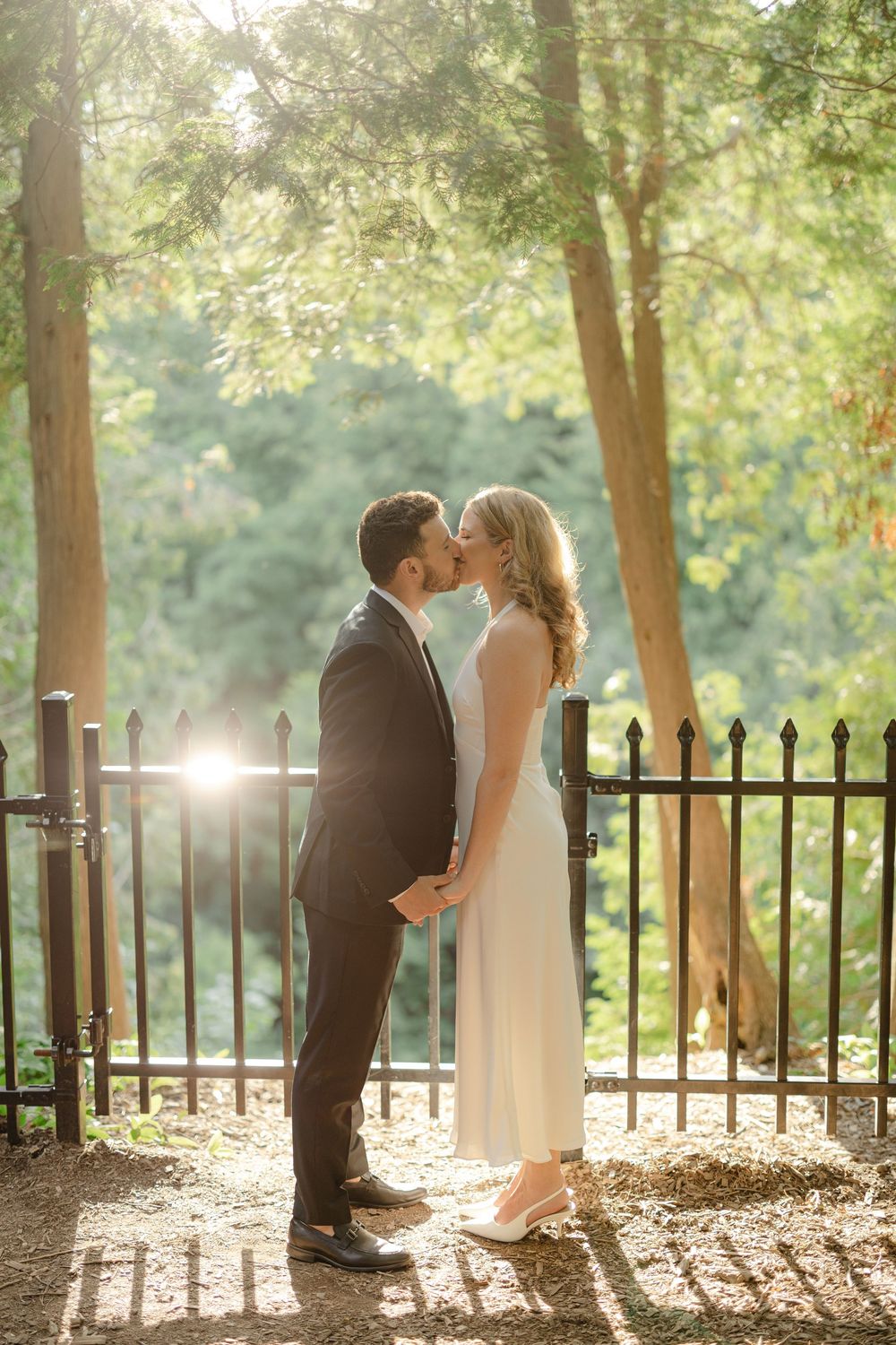 Bride in white dress and groom in black suit kissing while holding hands by an iron fence in sunlit woods.